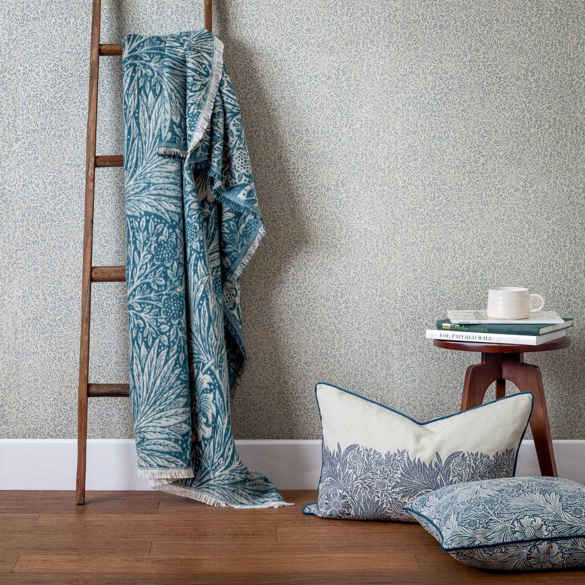 Marigold Jacquard Cotton Throw in indigo blue with a woven floral and leaf pattern, draped over a wooden ladder beside coordinating cushions and patterned wallpaper.