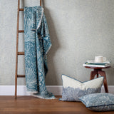 Marigold Jacquard Cotton Throw in indigo blue with a woven floral and leaf pattern, draped over a wooden ladder beside coordinating cushions and patterned wallpaper.