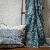 Marigold Jacquard Cotton Throw in indigo blue, draped with a fringed edge and woven botanical leaf pattern, shown beside matching cushions and a wooden ladder.