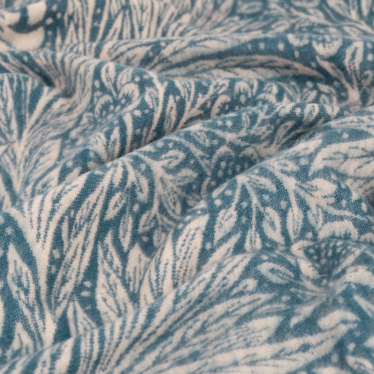 Marigold Jacquard Cotton Throw in indigo, close-up of the folded fabric showing a woven botanical leaf and floral pattern in teal-blue and cream.