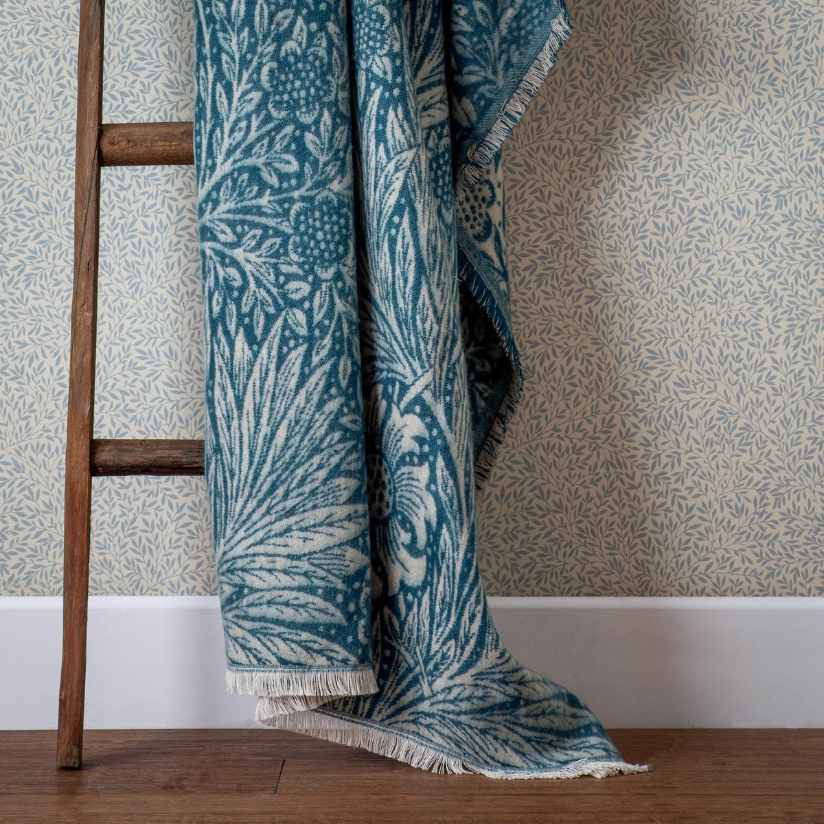 Marigold Jacquard Cotton Throw in indigo blue, draped over a wooden ladder against botanical-print wallpaper, showing the woven floral and leaf pattern with fringed edge.