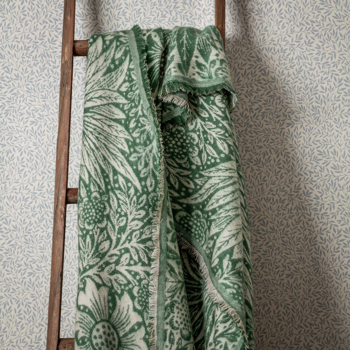 Marigold Jacquard Cotton Throw in Leaf Green draped over a wooden ladder, showing the reversible woven floral pattern in green and cream with a fringed edge.