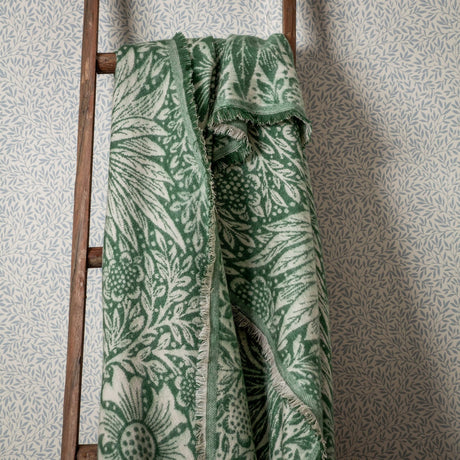 Marigold Jacquard Cotton Throw in Leaf Green draped over a wooden ladder, showing the reversible woven floral pattern in green and cream with a fringed edge.