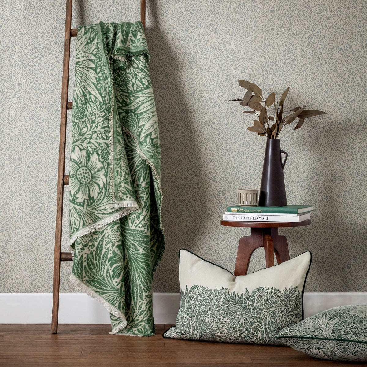 Marigold Jacquard Cotton Throw in Leaf Green draped over a wooden ladder, showing cream ground with green marigold foliage pattern and fringed edge, beside a coordinating cushion.