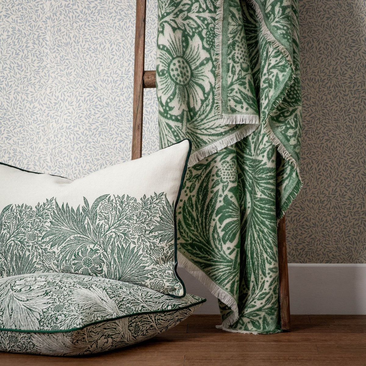 Marigold Jacquard Cotton Throw in Leaf Green draped with fringed edges, showing William Morris floral pattern in green on cream, styled with a matching cushion by a wooden ladder.