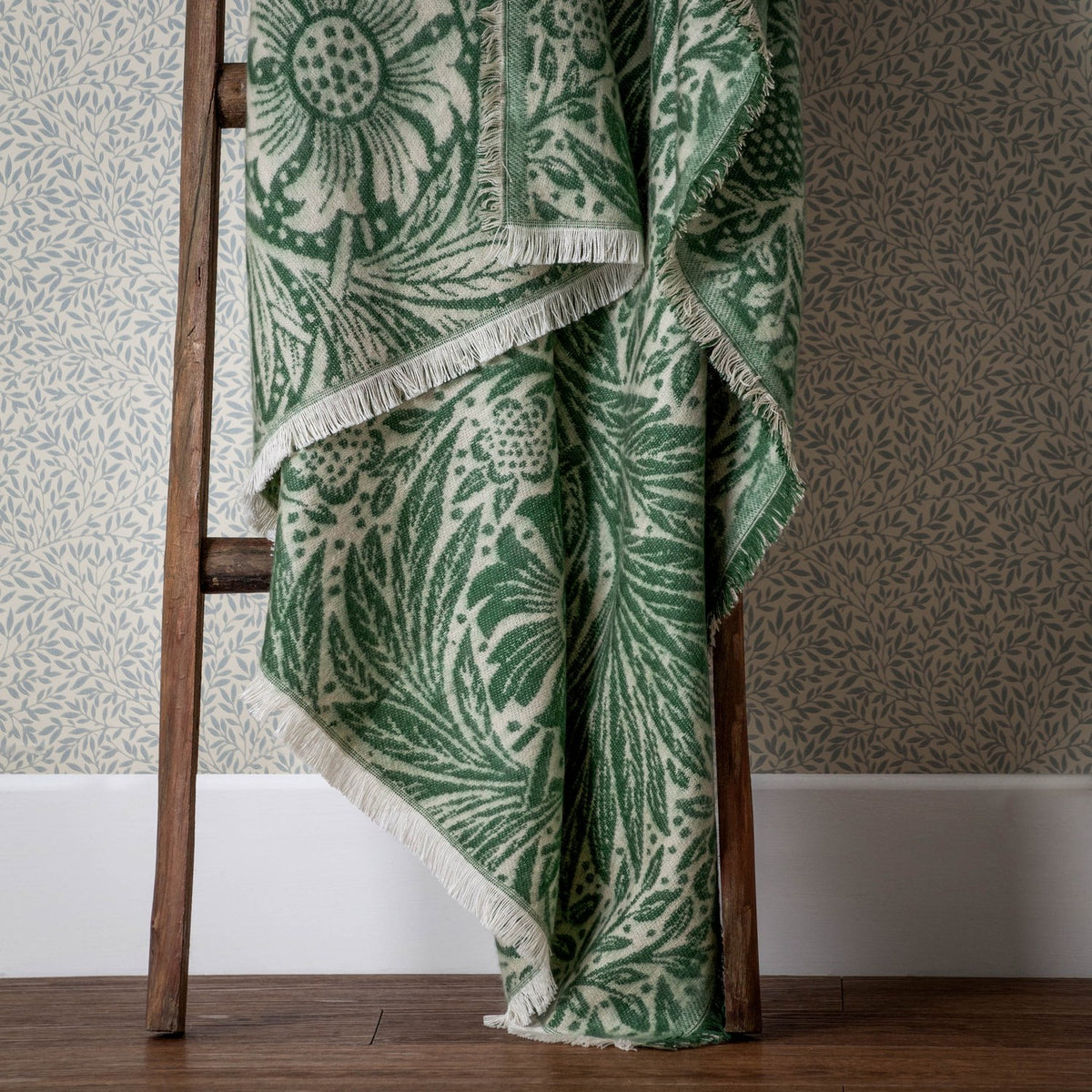 Marigold Jacquard Cotton Throw in leaf green, draped over a wooden ladder with fringed edges, showing William Morris floral pattern on cream ground.