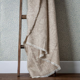 Marigold Jacquard Cotton Throw in stone, draped over a wooden ladder, showing its fringed edge and tonal leaf and floral pattern against blue floral wallpaper.