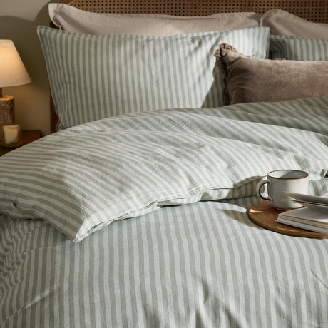 Marlow Stripe Cotton Printed Duvet Cover Set in Artichoke/Sage, shown on a bed with matching striped pillowcases, textured cushions, and a bedside lamp in a cosy bedroom setting.
