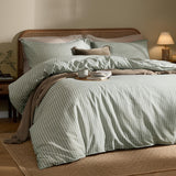 Marlow Stripe Cotton Printed Duvet Cover Set in Artichoke/Sage on a bed with matching pillowcases, sage-green stripes on off-white cotton, styled with cushions and a brown throw in a bedroom setting.
