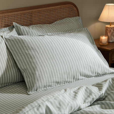 Marlow Stripe Printed Cotton Pillowcase Pair in artichoke/sage on a bed with rattan headboard, styled with striped duvet cover and bedside lamp.