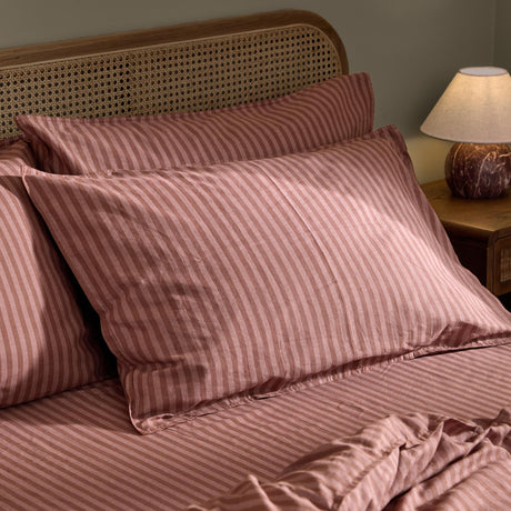 Marlow Stripe Printed Cotton Pillowcase Pair in Chocolate/Rosewood on a bedroom bed with a rattan headboard, showing the narrow vertical stripe print on a soft rose-brown ground.