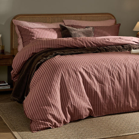 Bedroom scene showing the Marlow Stripe cotton pillowcase pair in rosewood on a bed with matching striped duvet, rattan headboard, and a dark brown throw blanket.