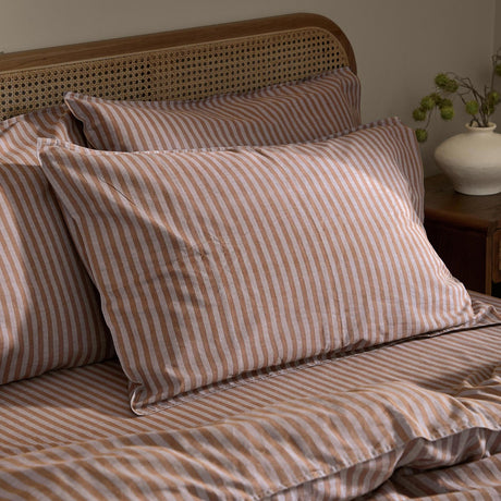 Marlow Stripe Printed Cotton Pillowcase Pair in cocoa, layered on a bed with matching striped duvet against a cane headboard in a bedroom setting.
