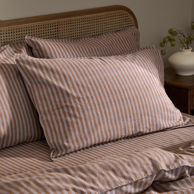 Marlow Stripe Printed Cotton Pillowcase Pair in cocoa, layered on a bed with matching striped duvet against a cane headboard in a bedroom setting.