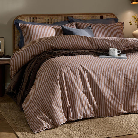 Bedroom scene with a made-up bed dressed in a Marlow Stripe cotton duvet set in cocoa and cream, layered with striped and plain vintage blue pillowcases against a rattan headboard.