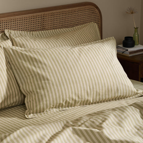 Marlow Stripe Printed Cotton Pillowcase Pair in Oregano/Shell, shown styled on a bed with a rattan headboard, featuring a soft green and cream stripe pattern on washed cotton fabric.