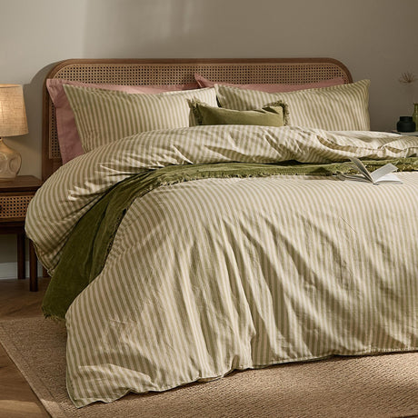 Marlow Stripe Printed Cotton Pillowcase Pair in Oregano/Shell on a bed with green and cream striped duvet, rattan headboard, and green velvet cushions.