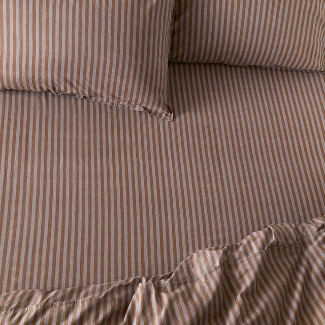 Marlow Stripe Printed Cotton Fitted Sheet in cocoa brown and vintage blue, showing a fine striped pattern on cotton fabric with matching pillowcases on a bed.