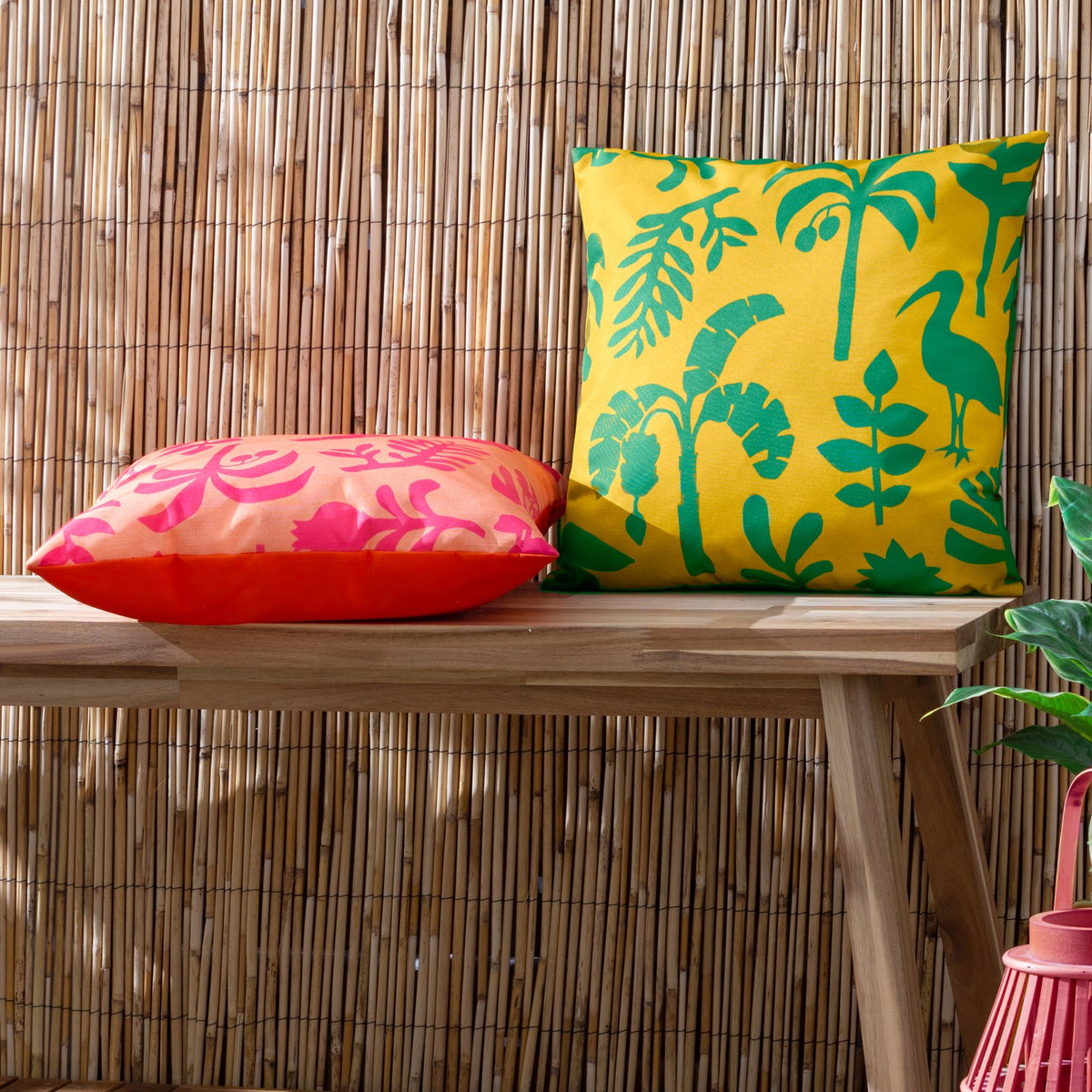 Marula Jungle polyester cushion in coral-orange with bold pink tropical print, shown beside a yellow botanical cushion on a wooden bench against a bamboo fence.