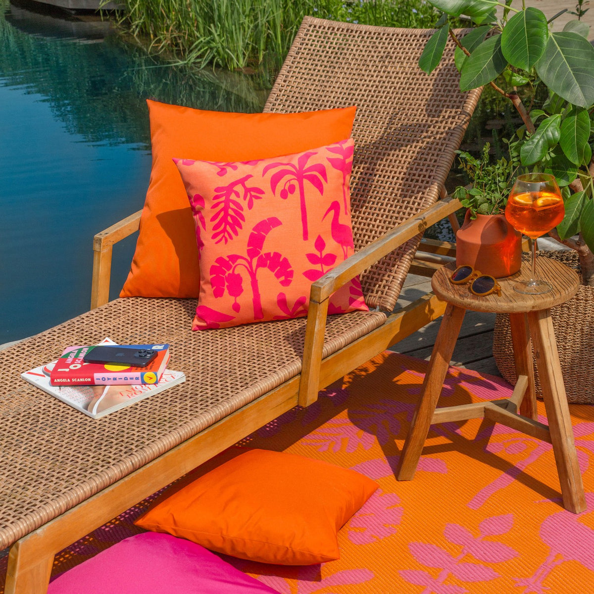 Marula Jungle polyester cushion in coral-orange with a bold pink tropical print, styled on a rattan lounger beside a plain orange cushion and outdoor rug.