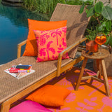 Marula Jungle polyester cushion in coral-orange with a bold pink tropical print, styled on a rattan lounger beside a plain orange cushion and outdoor rug.