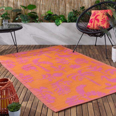 Marula Reversible Jungle Rug in orange and pink with tropical leaf and bird patterns, shown on a wooden deck beside a chair and plants.