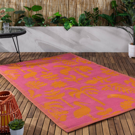 Marula Reversible Jungle Rug in pink and orange with tropical leaf and bird silhouette pattern, laid on a wooden deck beside potted plants and a bamboo fence.