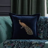Mayura Animal Velvet Cushion in midnight blue, embroidered peacock design with gold and beaded tail detail, styled on a teal tufted sofa with patterned throw.