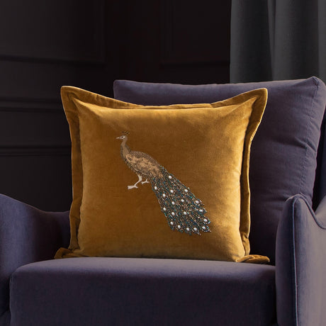 Mayura mustard-gold velvet cushion with hand-embroidered beaded peacock design, shown on a dark blue armchair.