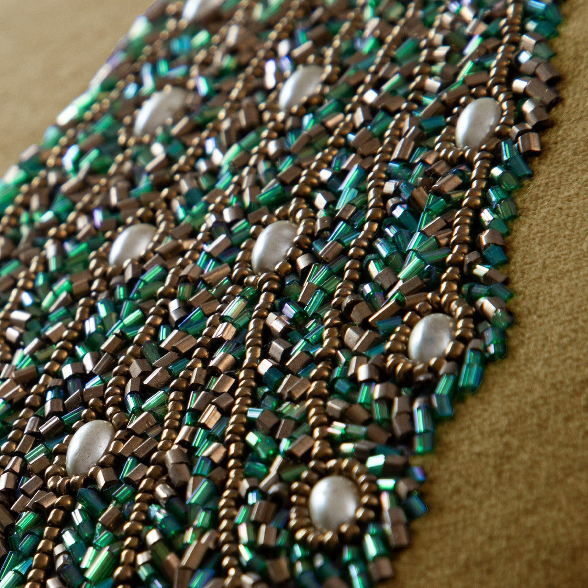 Close-up of the Mayura velvet cushion's hand-embroidered peacock detail, showing green and bronze beadwork with pearl accents on a mustard-gold velvet ground.