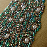 Close-up of the Mayura velvet cushion's hand-embroidered peacock detail, showing green and bronze beadwork with pearl accents on a mustard-gold velvet ground.