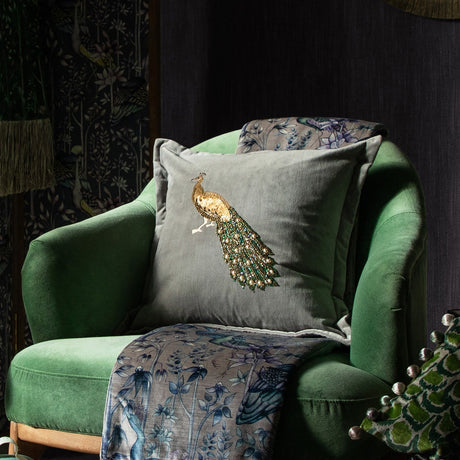 Mayura Animal Velvet Cushion in Steel grey with beaded peacock design, styled on a green velvet armchair alongside a patterned throw.