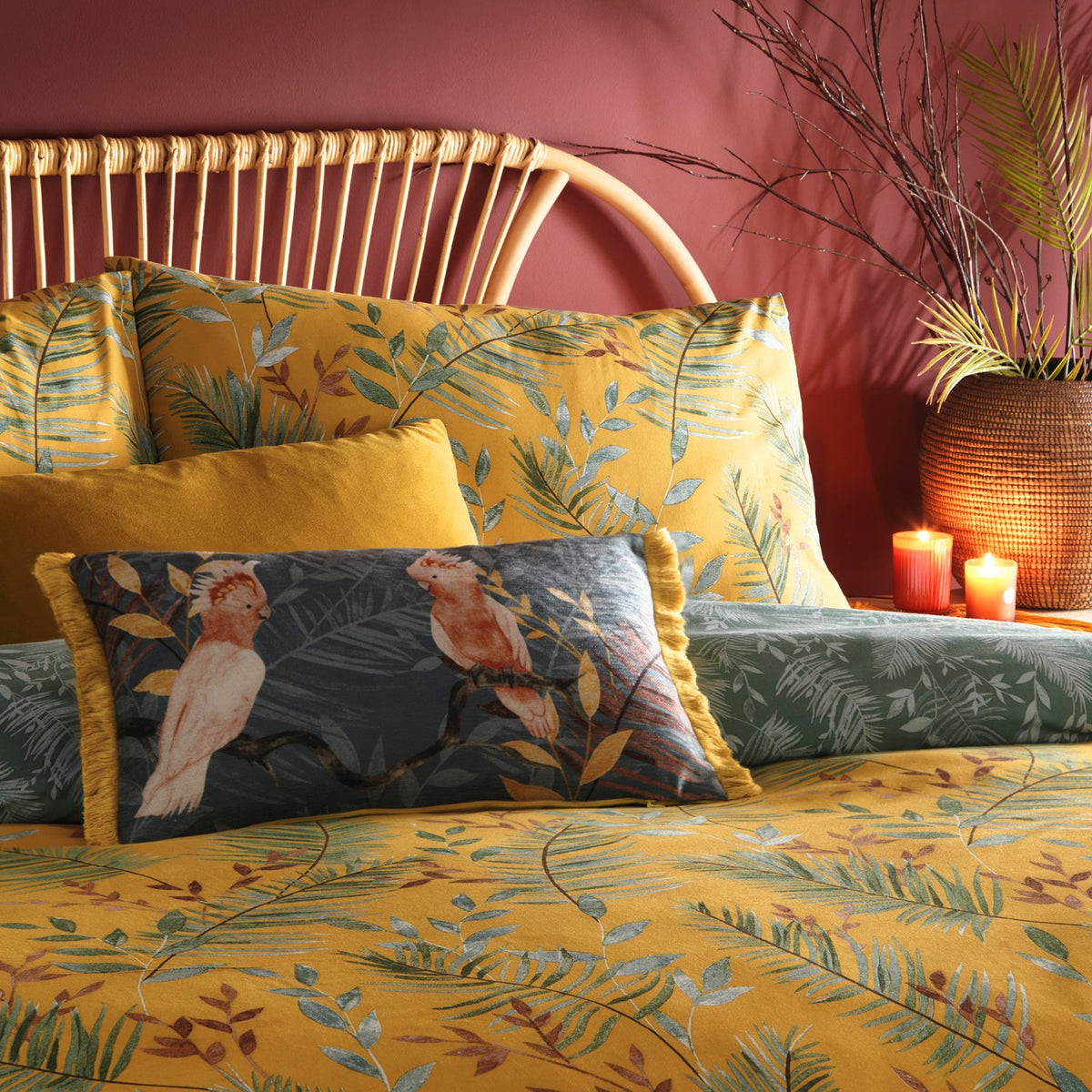 Mazari Printed Jungle Duvet Cover Set in mustard on a bedroom bed, showing palm frond and leaf patterns with matching pillowcases and a bird-print bolster cushion.