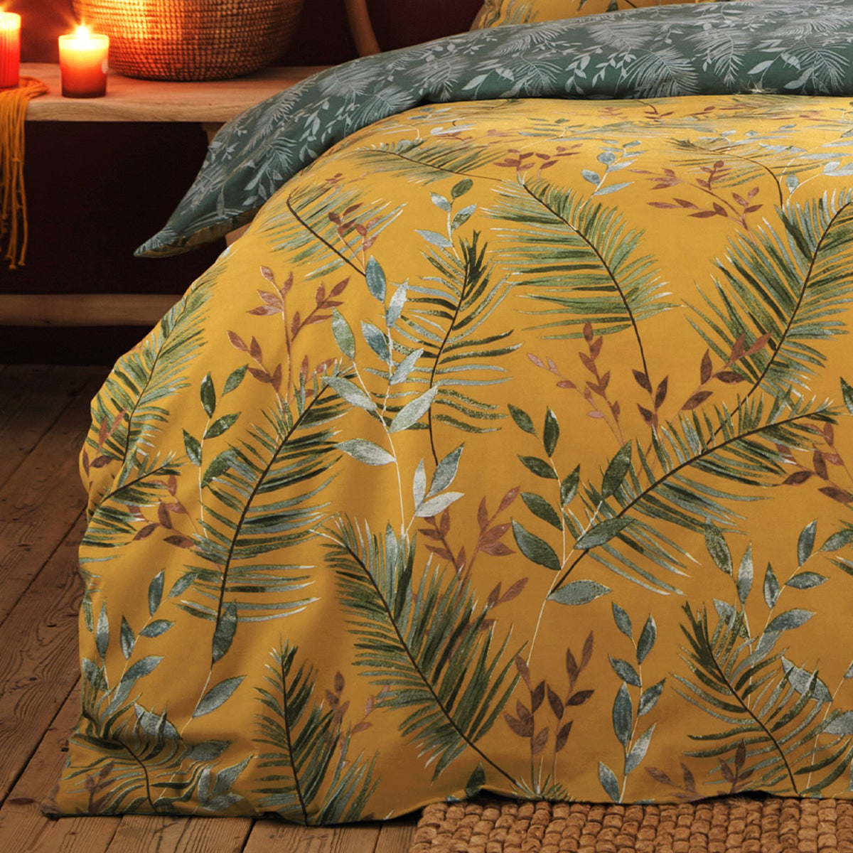 Mazari Printed Jungle Duvet Cover Set in mustard yellow with green palm fronds and rust leaves, shown on a bed with a matching green leaf-print pillowcase in a warm bedroom.