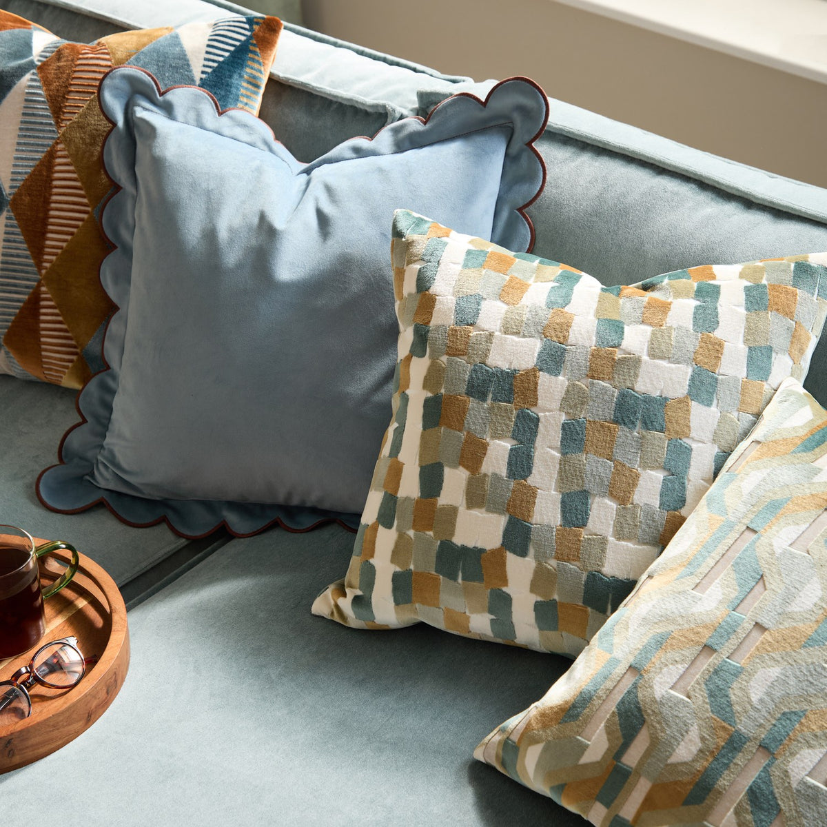 Metro Blocks Jacquard Velvet Cushion in green shown on a light blue sofa, featuring an abstract woven block pattern in teal, ochre and cream with a square scatter-cushion shape.