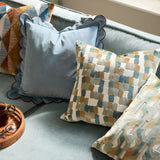 Metro Blocks Jacquard Velvet Cushion in green shown on a light blue sofa, featuring an abstract woven block pattern in teal, ochre and cream with a square scatter-cushion shape.