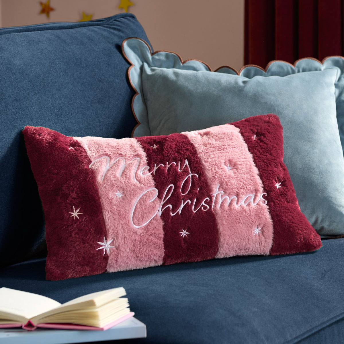 Rectangular faux fur cushion in burgundy and pink stripes with embroidered "Merry Christmas" text and stars, shown on a blue sofa with light blue scalloped cushions.