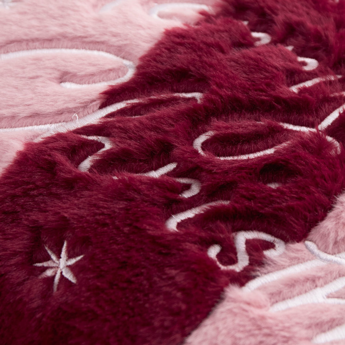Close-up of the Merry Christmas Plain Faux Fur Cushion in burgundy and pink, showing plush pile fabric with embroidered lettering and star details.