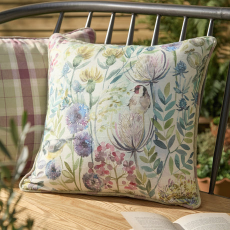 Morning Chorus Linen Cotton Cushion in Elderberry, square with hand-painted goldfinch, songbird and floral design, shown on an outdoor bench beside a check-patterned cushion.