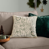 Merry Christmas Embroidered Velvet Cushion in ecru with dark green festive motifs and gold piping, shown on a sofa beside a plain green velvet cushion.