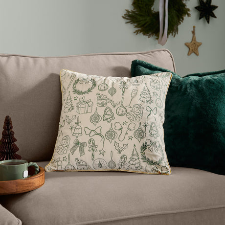 Merry Christmas Embroidered Velvet Cushion in ecru with dark green festive motifs and gold piping, shown on a sofa beside a plain green velvet cushion.