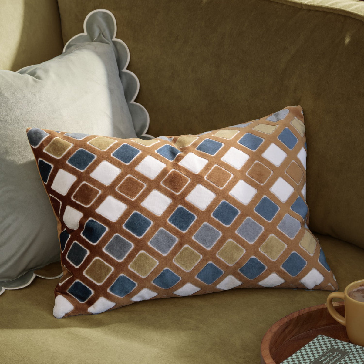 Metro Diamond Jacquard Velvet Cushion in gold and blue, rectangular with a rust-toned diamond pattern, shown on an olive-green sofa beside a scalloped grey pillow.
