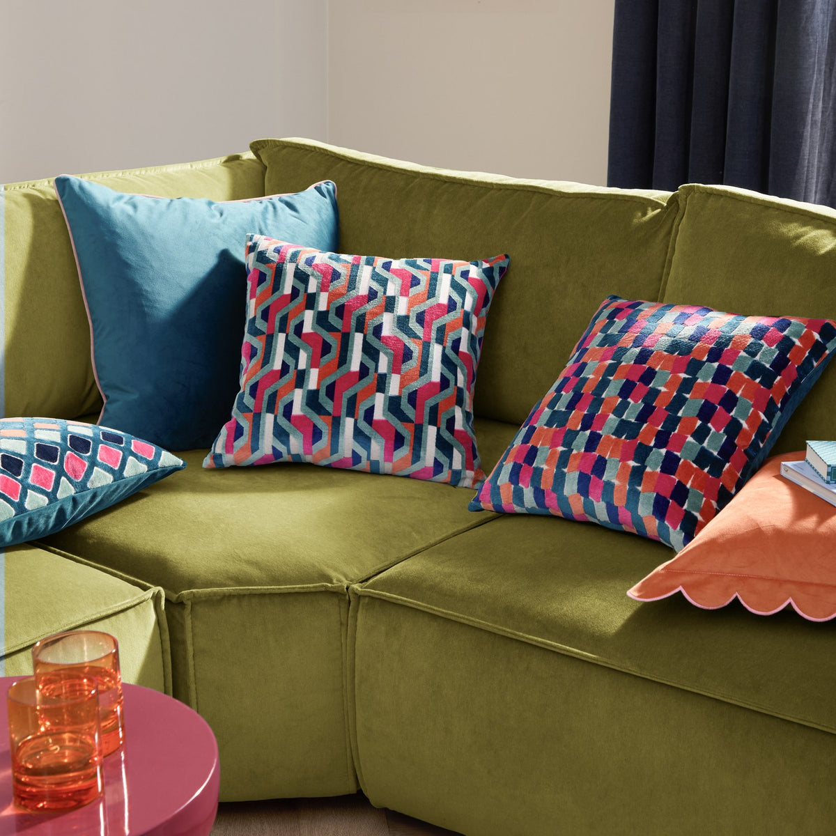 Metro Diamond jacquard velvet cushion in a multicoloured diamond grid pattern with teal, pink, orange and navy tones, styled on an olive green sofa alongside patterned and plain teal cushions.