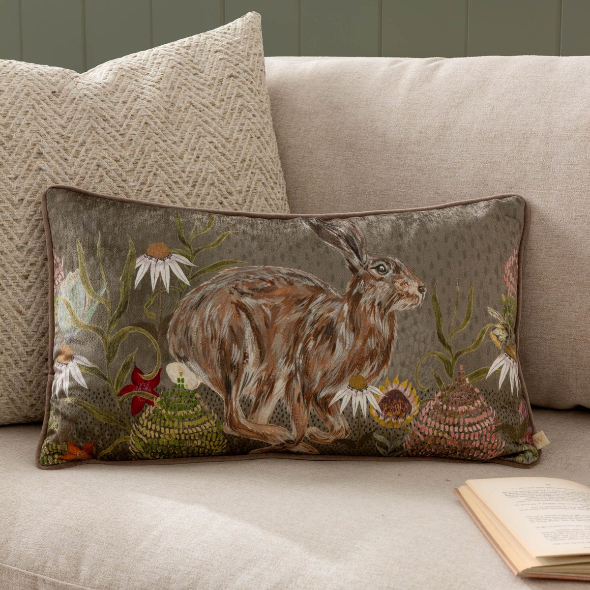 Rectangular sage green chenille cushion with printed hare and wildflower design, styled on a beige sofa beside a textured cream cushion.