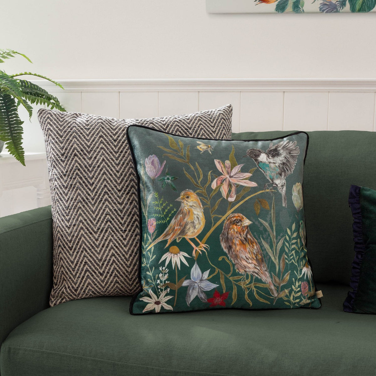 Meadow Bloom Animal Chenille Cushion in teal, printed with hand-painted birds, wildflowers and foliage, shown on a green sofa beside a chevron-patterned cushion.