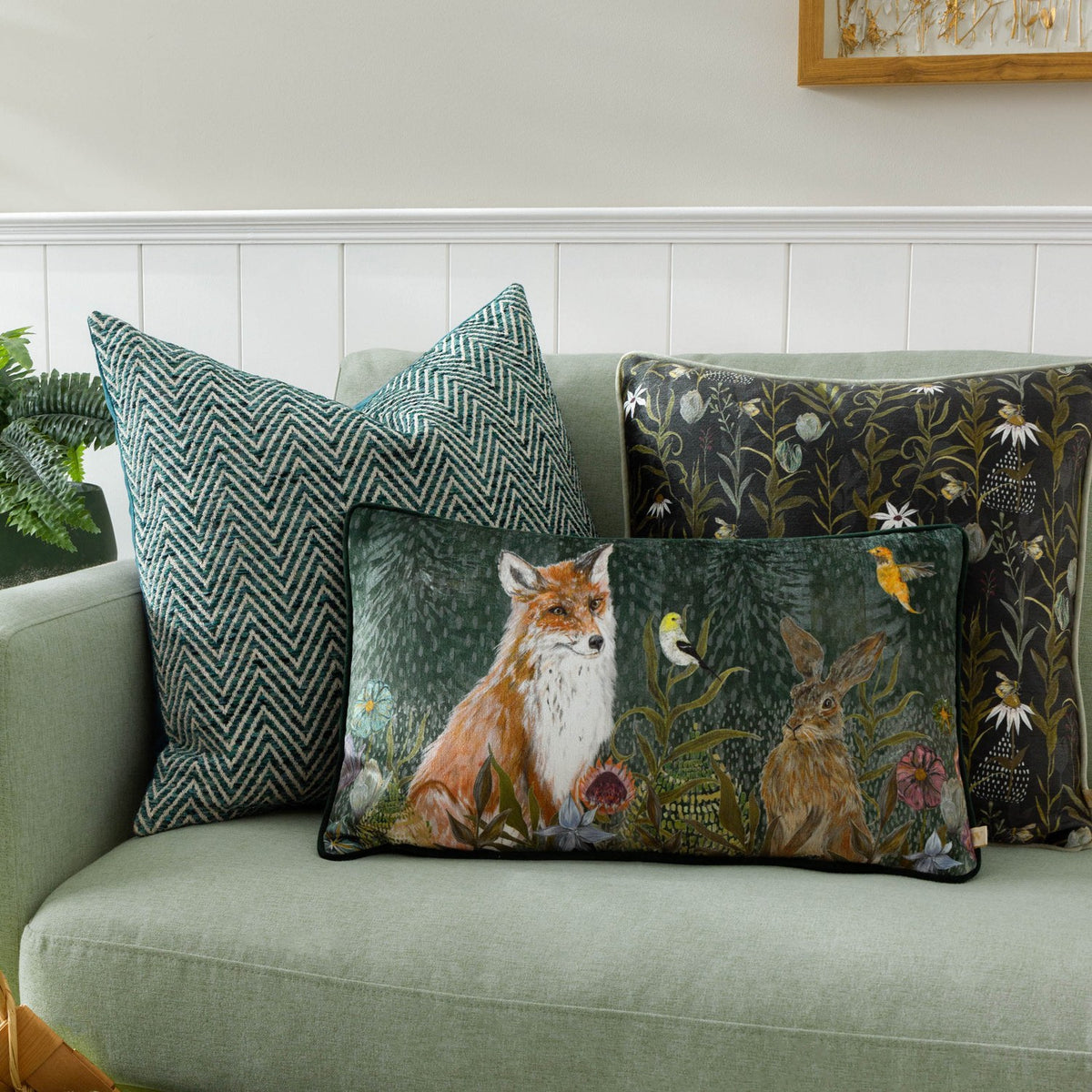 Rectangular fox and hare woodland cushion on a green sofa, styled with a black floral chenille cushion and a green herringbone cushion against a white panelled wall.