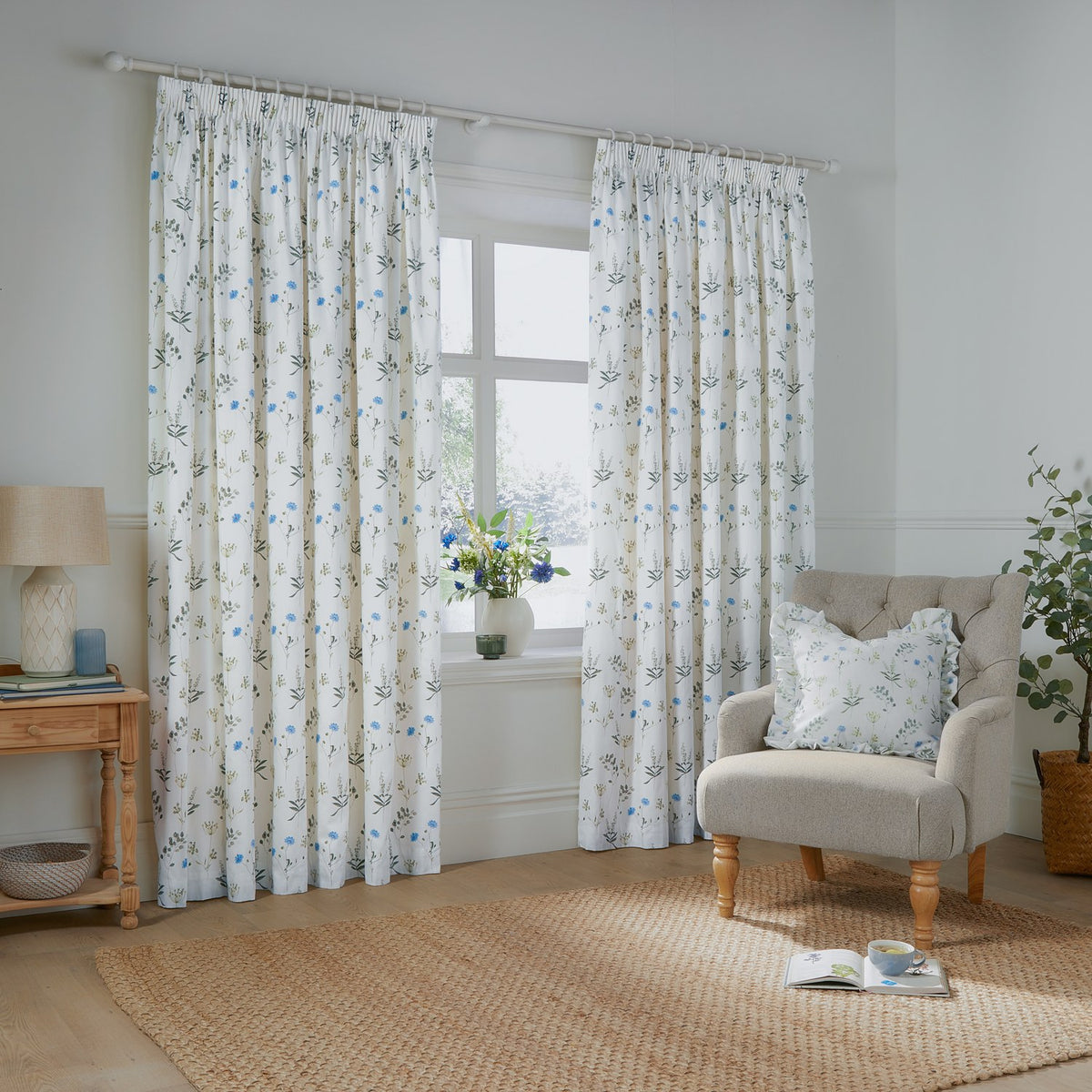 Meadow Floral Cotton Pencil Pleat Curtains in white with cornflower-blue and green wildflower print, shown pleated on a white pole in a living room beside an armchair.