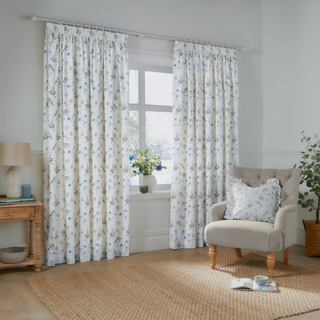 Meadow Floral Cotton Pencil Pleat Curtains in white with cornflower-blue and green wildflower print, shown pleated on a white pole in a living room beside an armchair.