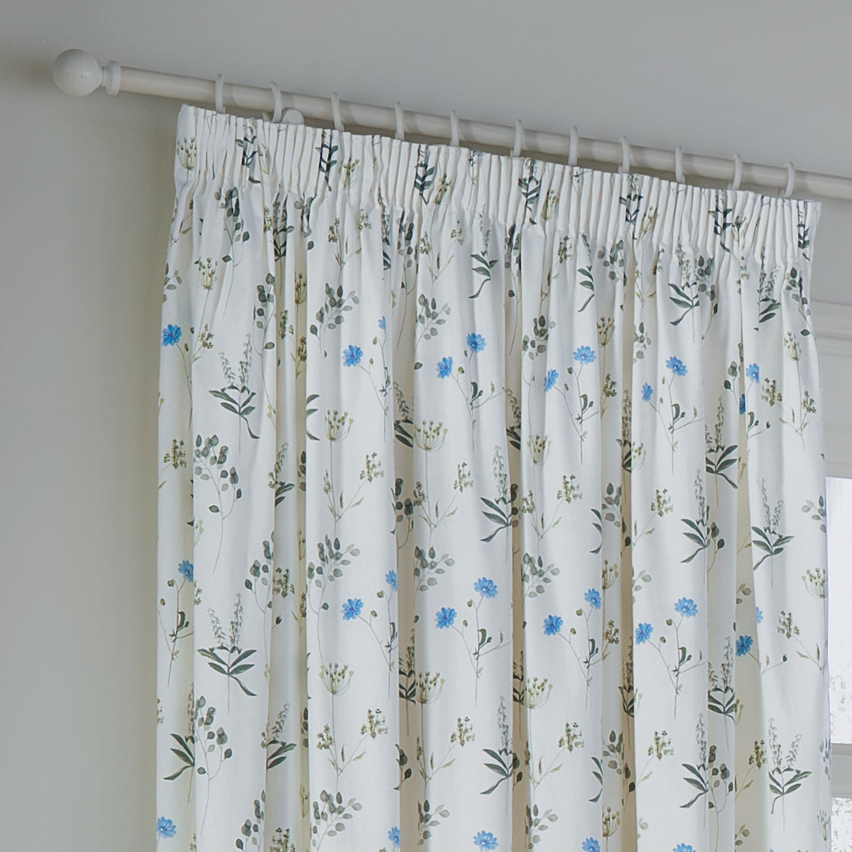 Pencil pleat curtain in white cotton Panama with a scattered meadow print of cornflower-blue blooms and green leaves, shown on a white pole against a light grey wall.