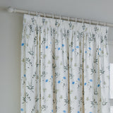 Pencil pleat curtain in white cotton Panama with a scattered meadow print of cornflower-blue blooms and green leaves, shown on a white pole against a light grey wall.